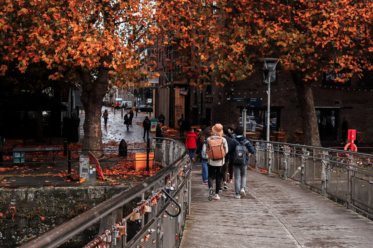 People with backpacks cross a footbridge beneath trees in full orange autumn colour, padlocks along the railing.