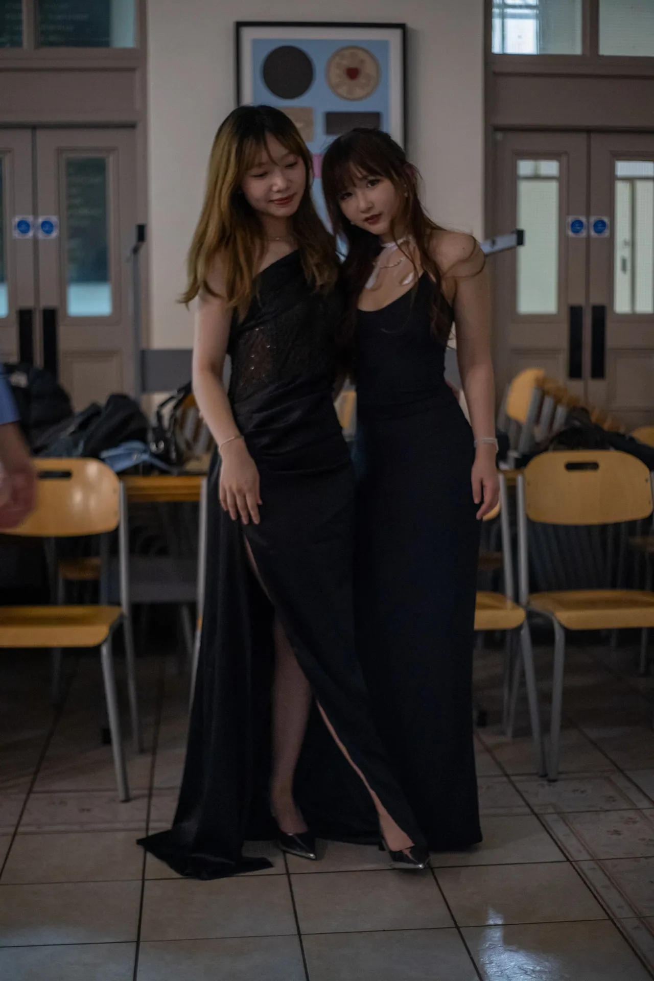 Two young women in long black dresses pose together in a hall, stacked chairs behind them.