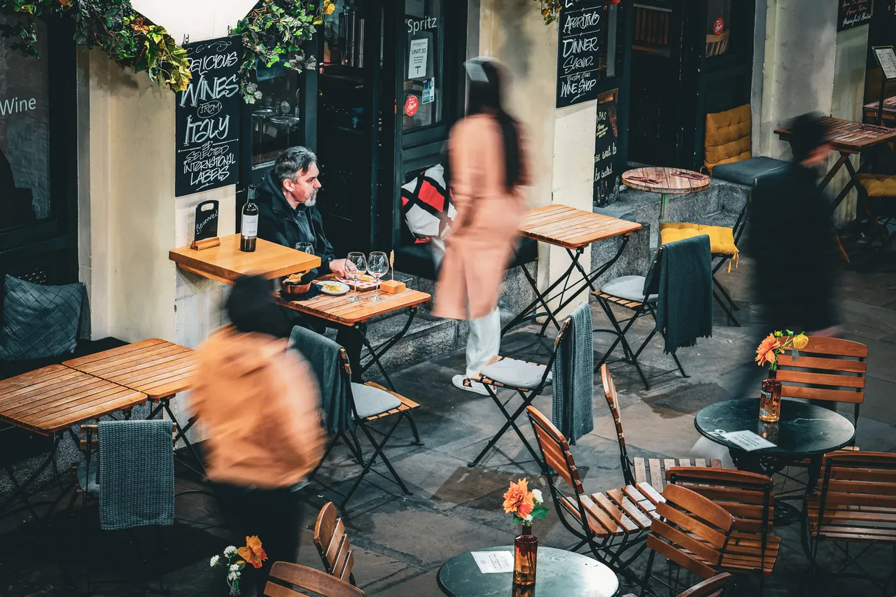 An outdoor café terrace in motion — a man sits still at his table while staff and passers-by blur around him.