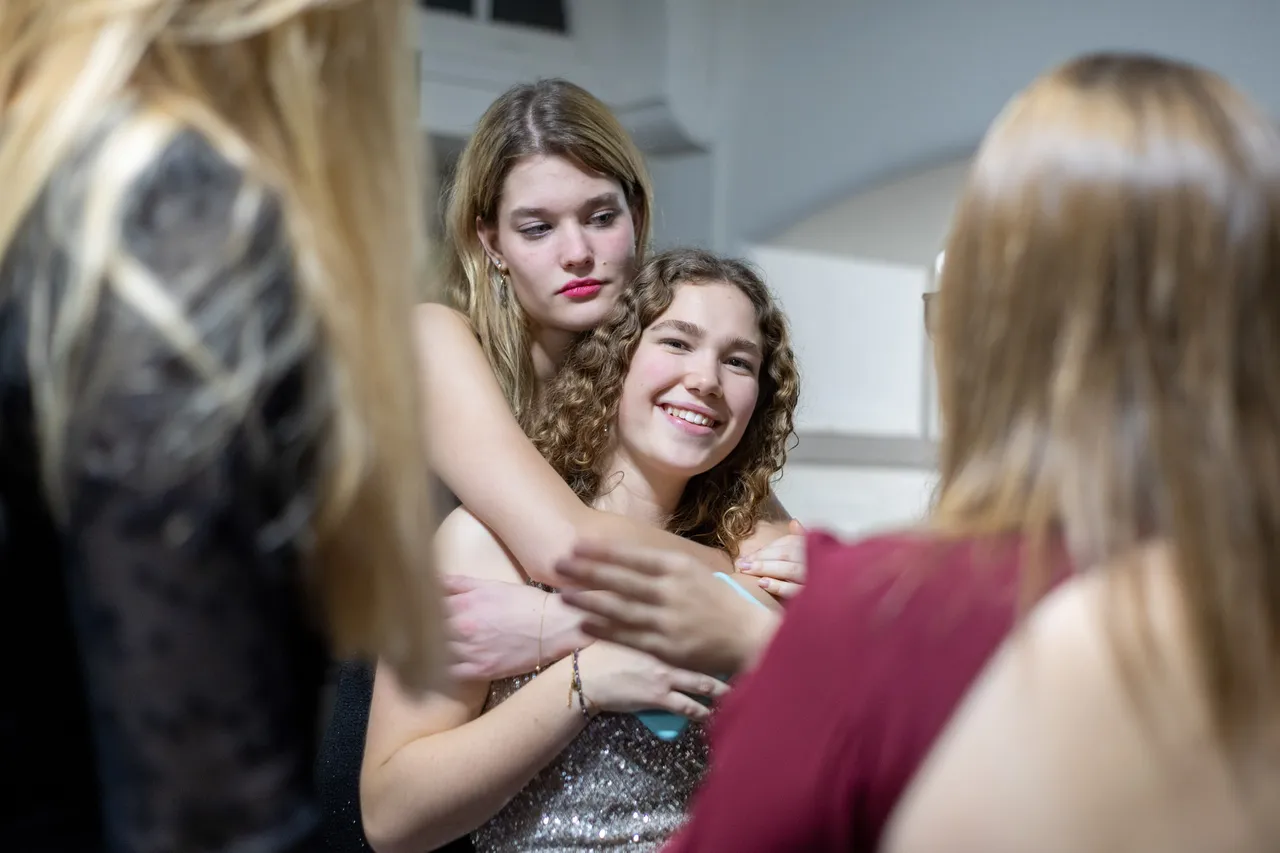 At a party, a young woman in sequins is pulled into a hug by a friend, framed between the shoulders of other guests.