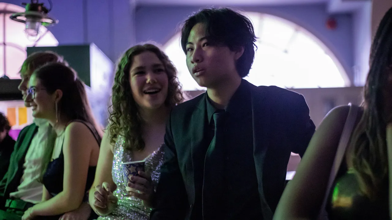 A young man in a dark suit sits among friends at a party, green and violet light washing across the room.