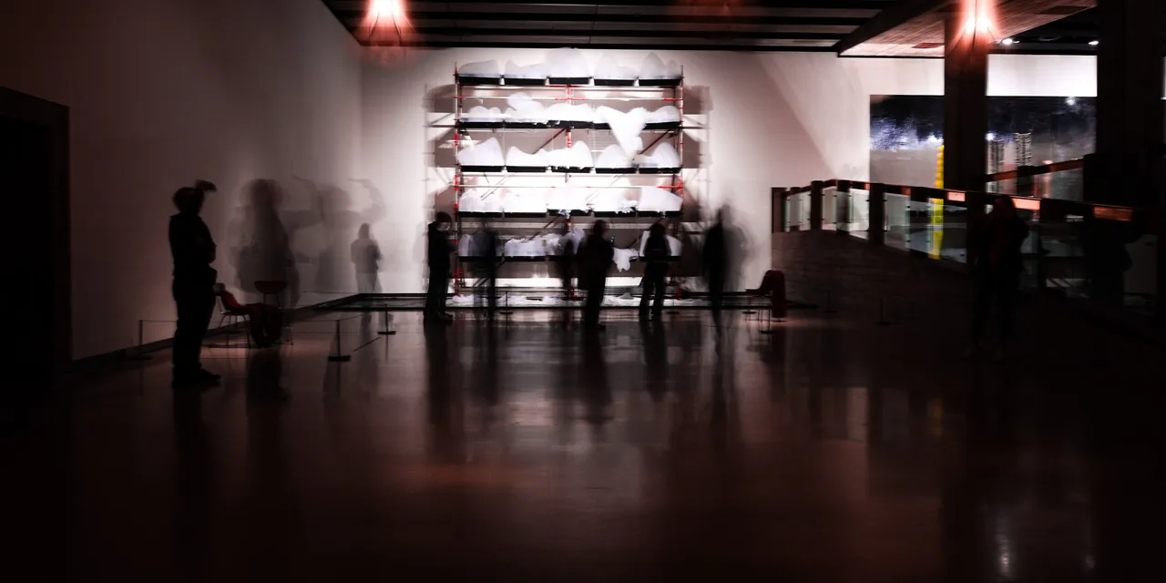 Visitors reduced to shadows and ghosted motion in a darkened gallery, the floor reflecting a lit display wall.