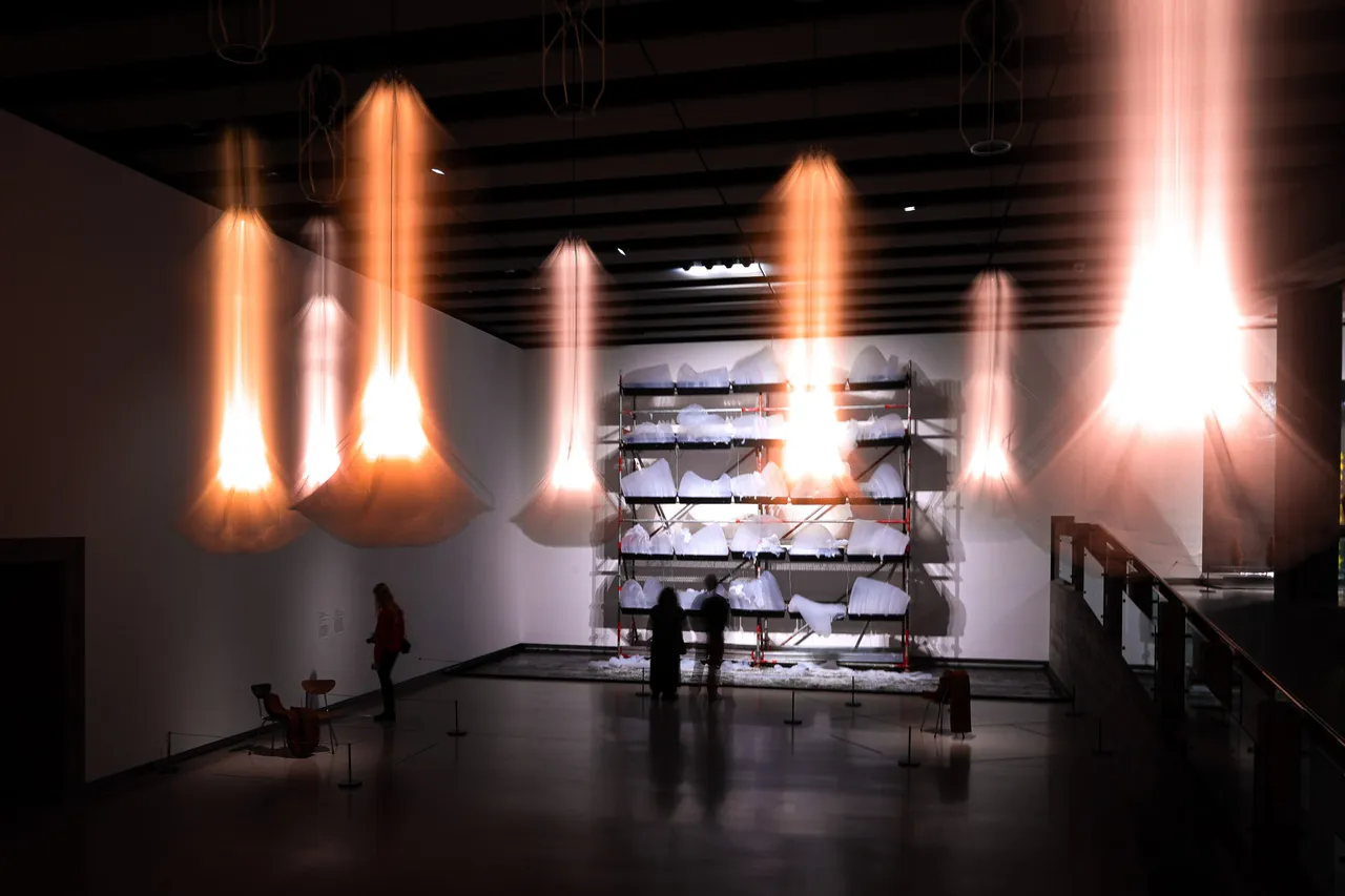 Tall glowing lamps hang in a dark gallery, blurred into streaks of light above a few visitors and a wall of white sculptures.