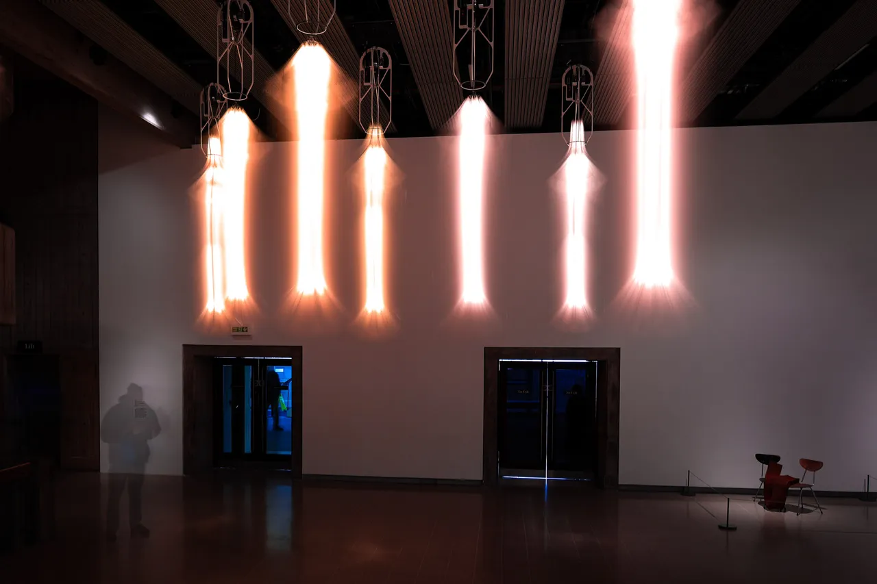 A row of hanging lamps smeared into vertical flares of light above two doorways in a white gallery wall.
