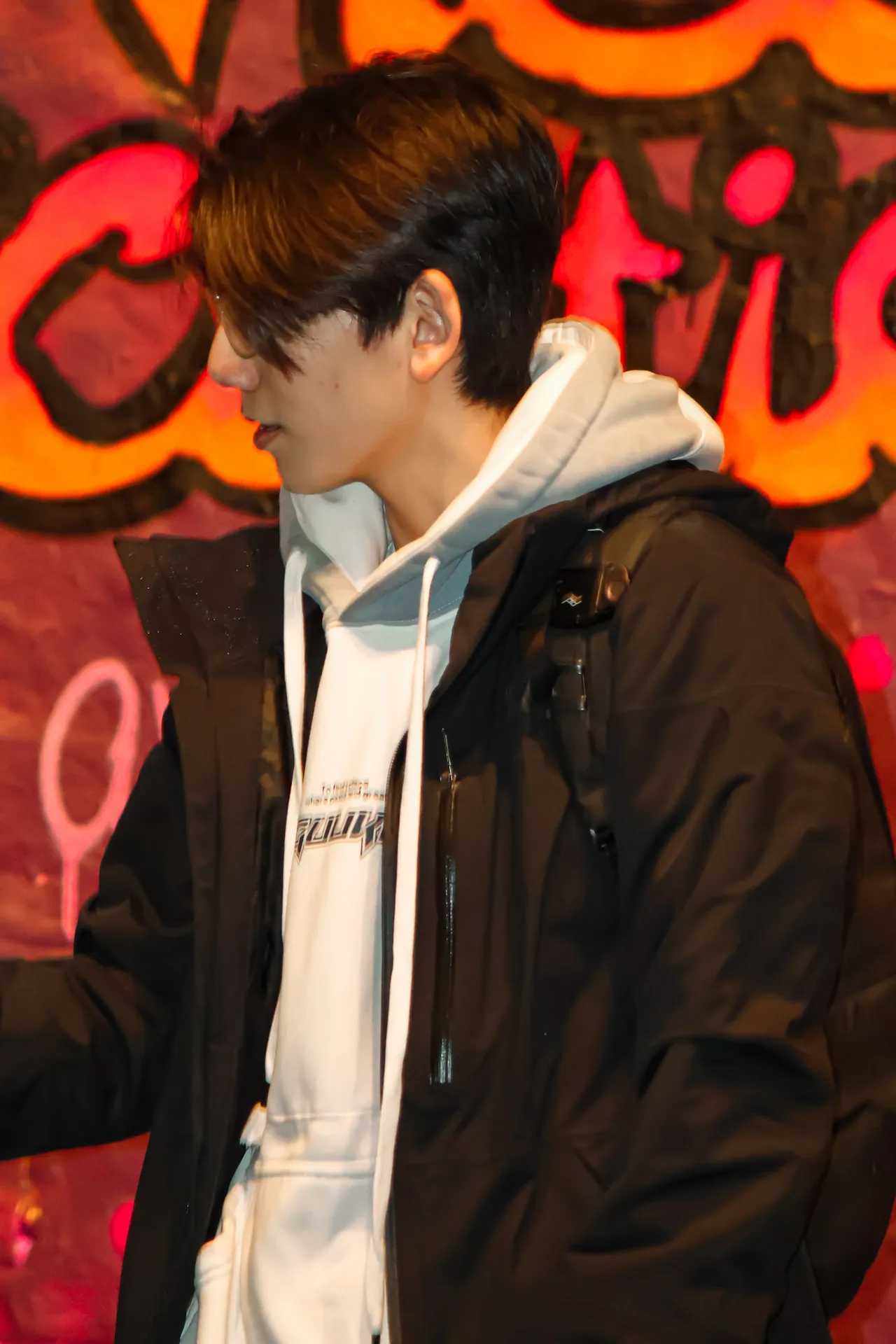 A young man in a white hoodie and dark jacket turns away against a wall of red and orange graffiti.