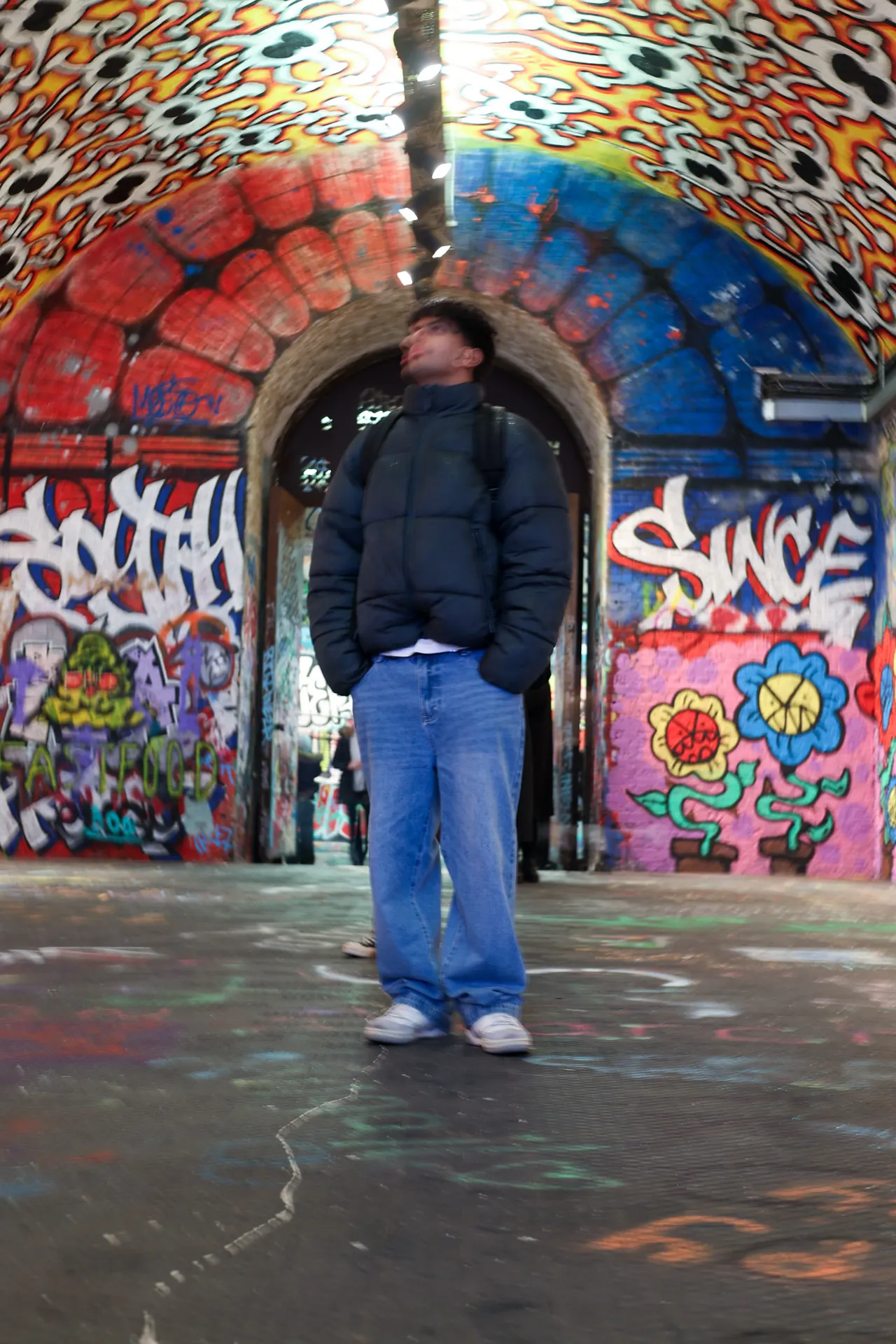 A man in a black puffer jacket and wide jeans stands in a graffiti-covered tunnel, looking up at the painted arch.