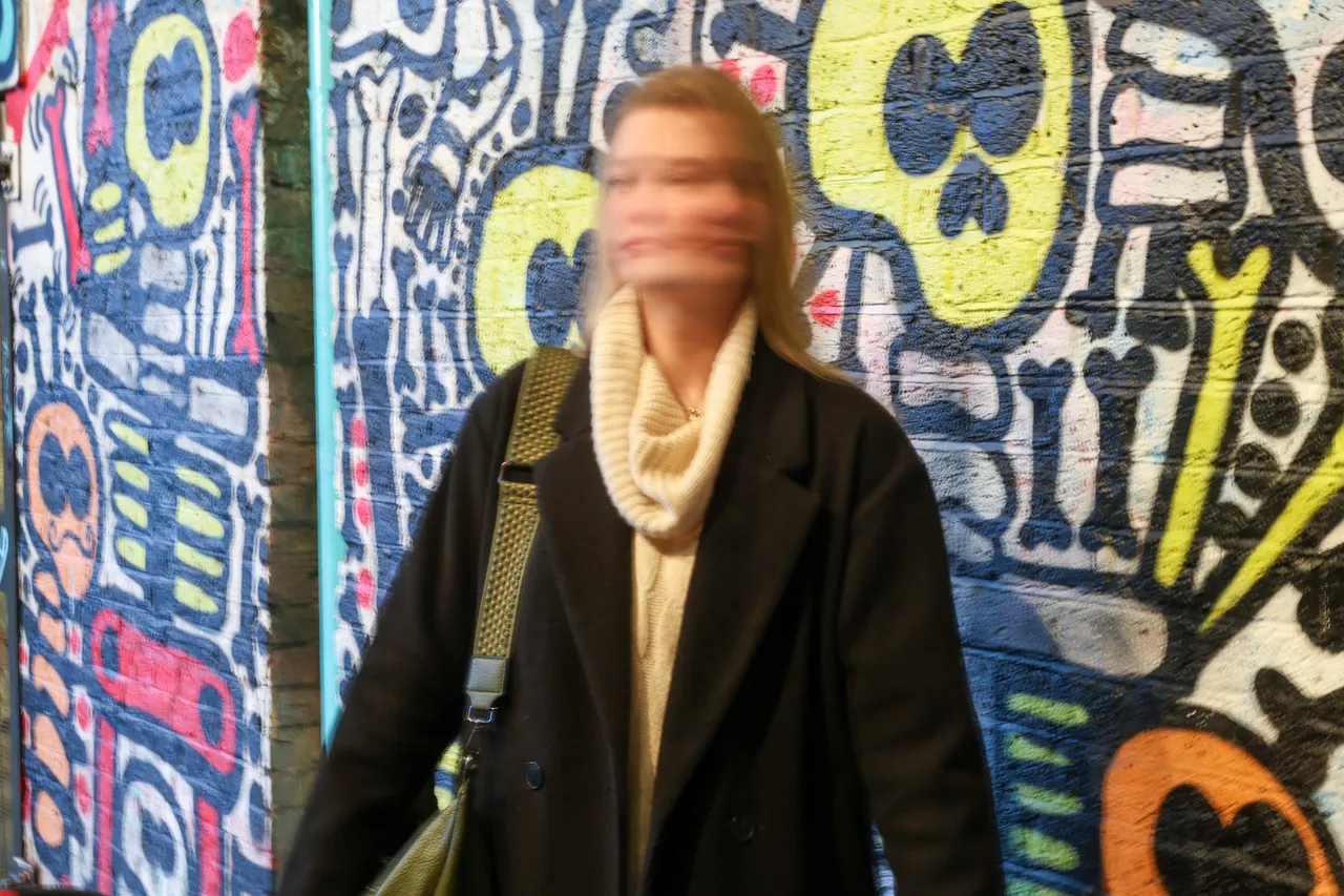 A woman in a dark coat and cream scarf moves past a mural of cartoon skulls, her face blurred by motion.