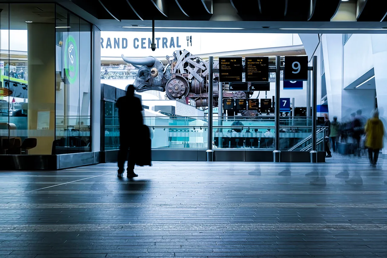 A lone figure stands in a bright station concourse beneath a large “Grand Central” sign as other travellers blur past.