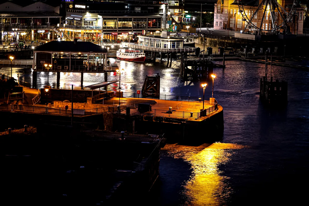 Night over a harbour — moored boats and waterfront bars glow orange, their lights spilling across black water.