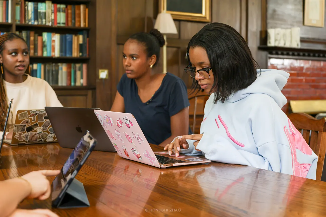 Students work on laptops around a polished table in a wood-panelled library.