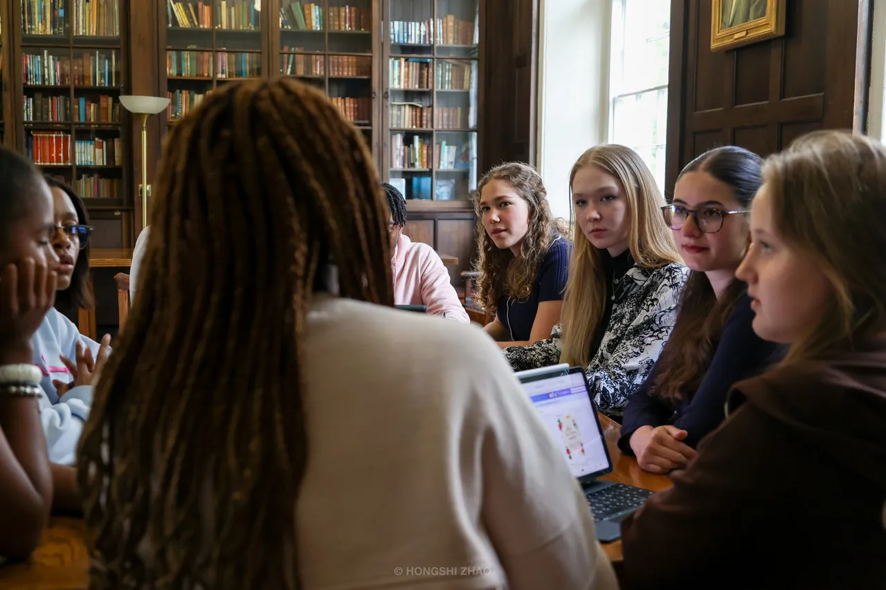 Seen over a shoulder, a group of students listen around a library table lined with bookshelves.