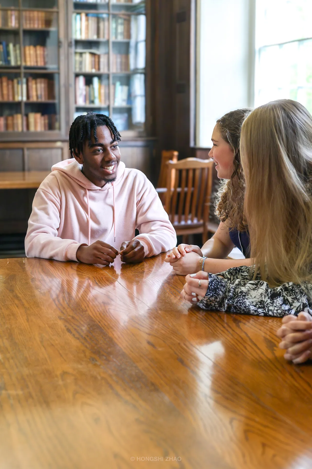 A student in a pink hoodie smiles across a long, reflective library table.