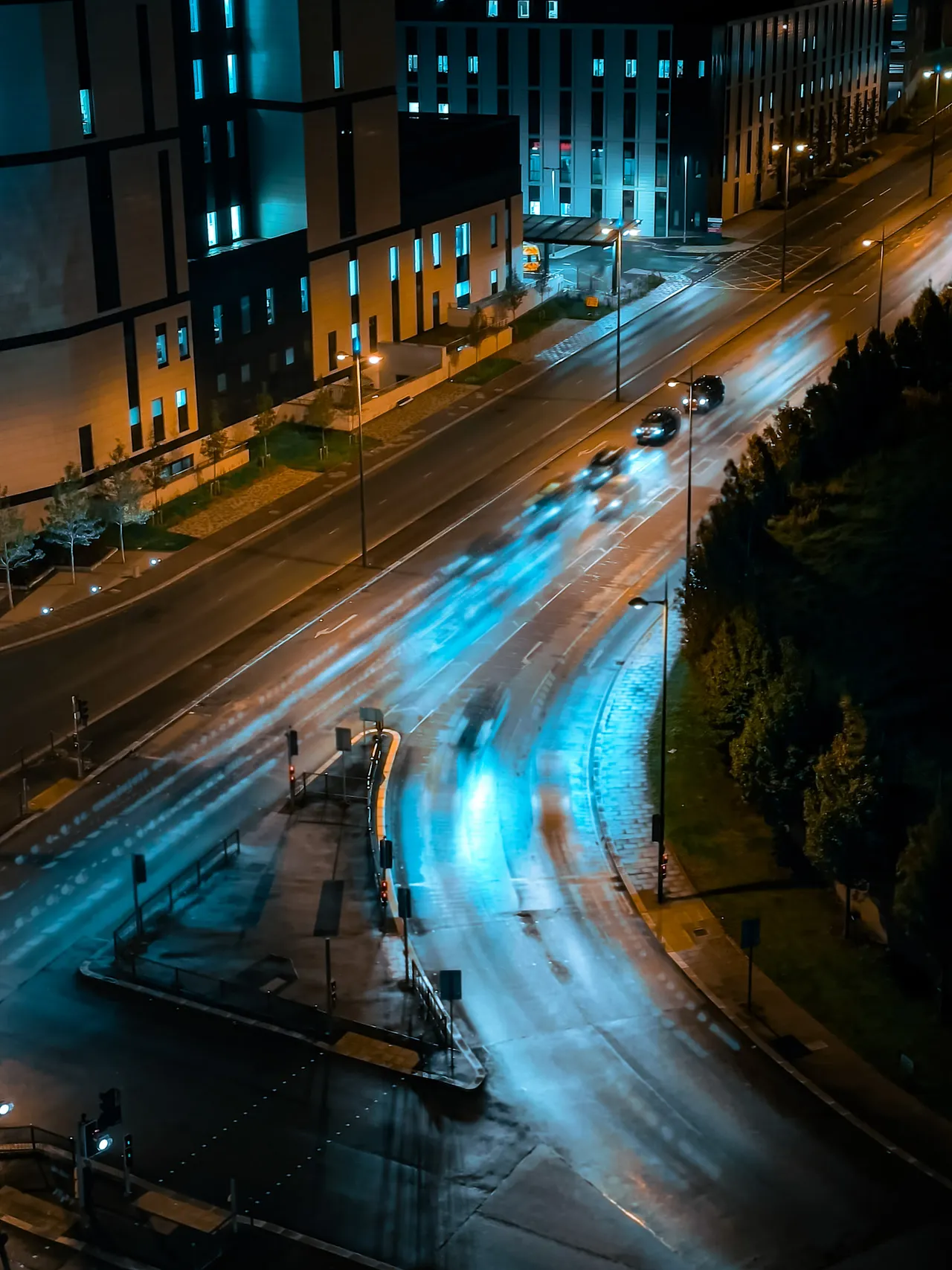 From above at night, headlights trace blue and white trails around a curving city road beside dark trees and apartment blocks.