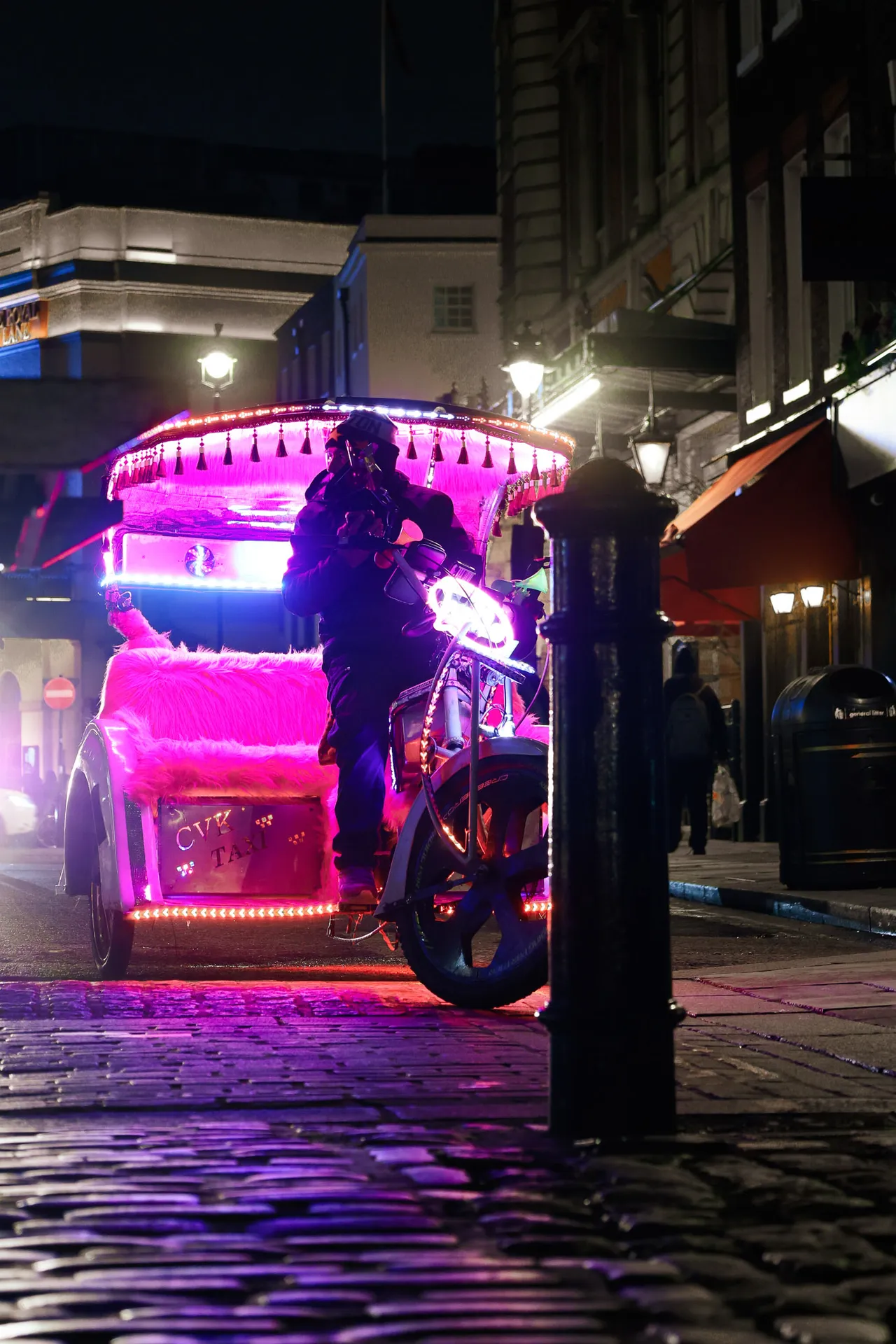 A rickshaw lit in hot-pink neon waits on a cobbled street at night.