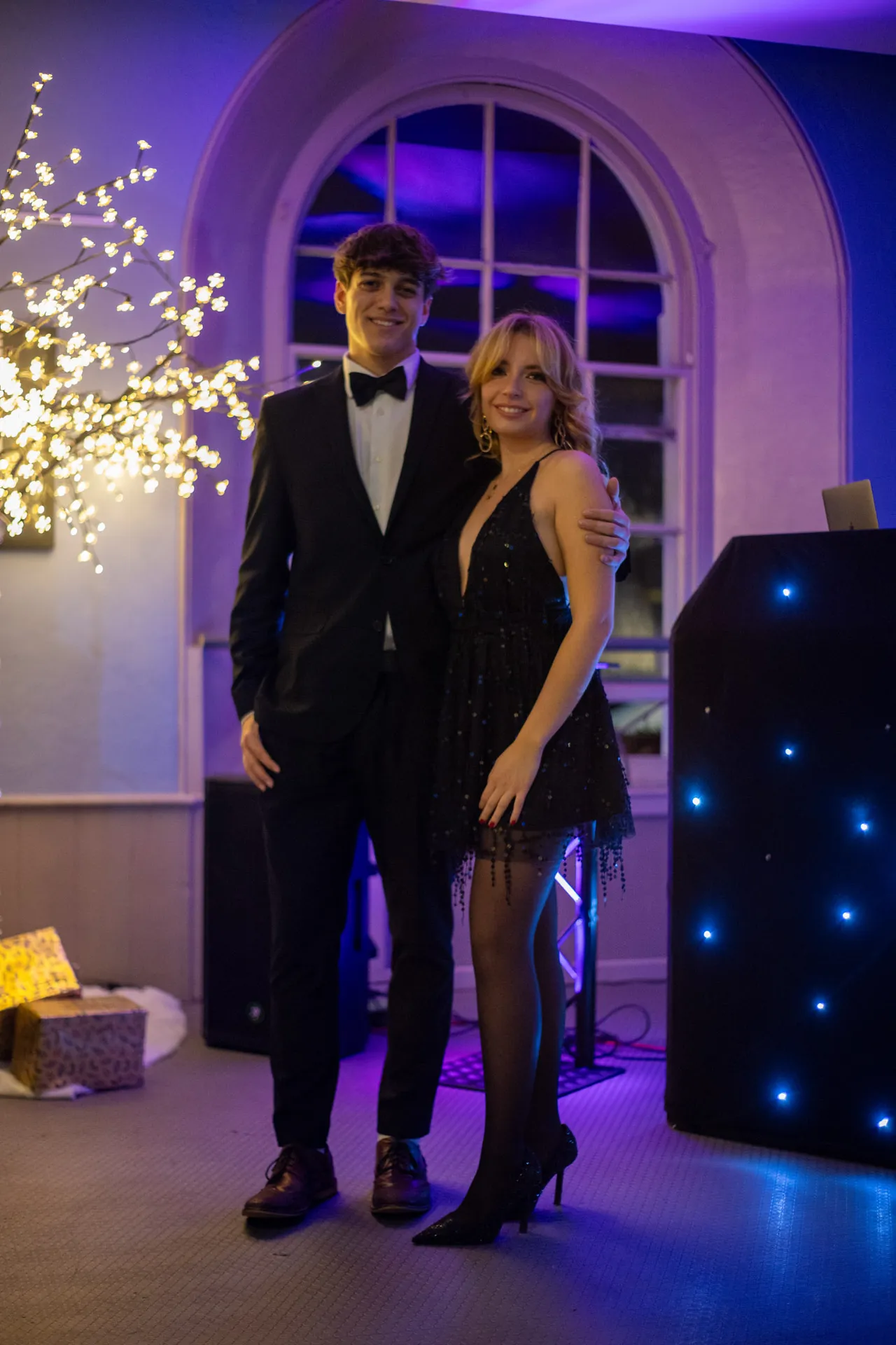 A couple in formal wear stand side by side beneath an arched window, lit in blue and violet.