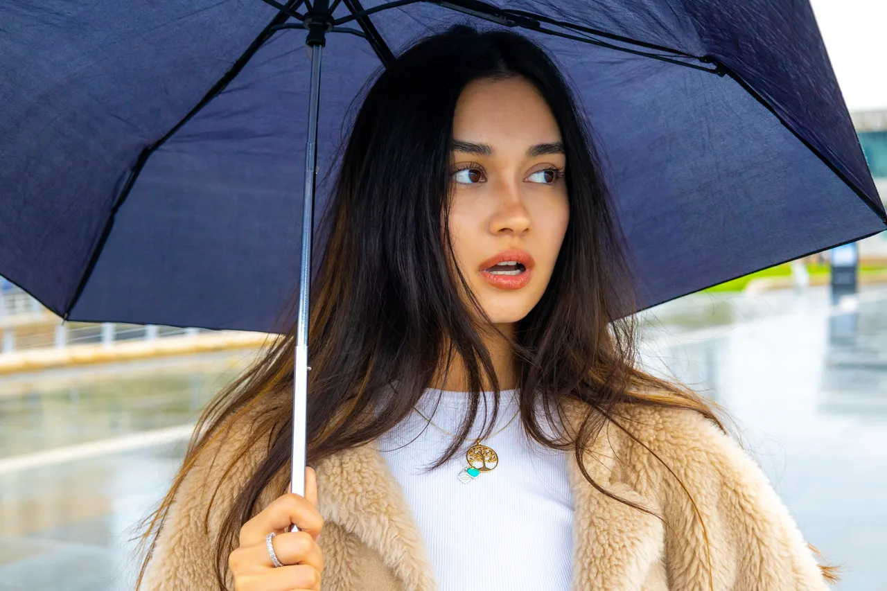 A young woman with long dark hair shelters under a navy umbrella on a rain-wet waterfront, glancing to one side.