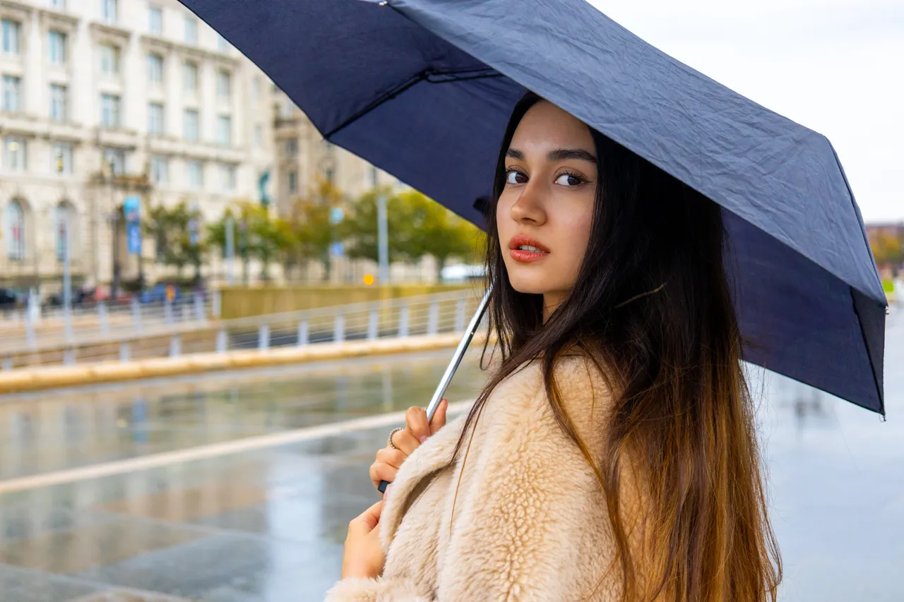 The same woman looks back over her shoulder beneath her umbrella, a grand stone building and wet promenade behind her.
