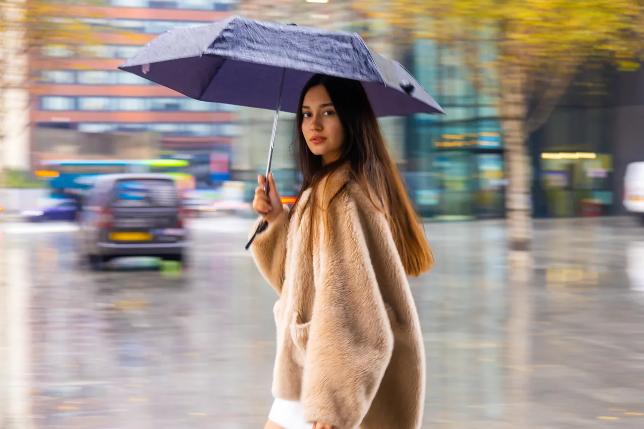 Panned in the rain — the woman in a camel coat stays sharp under her umbrella while traffic and city lights streak past behind her.