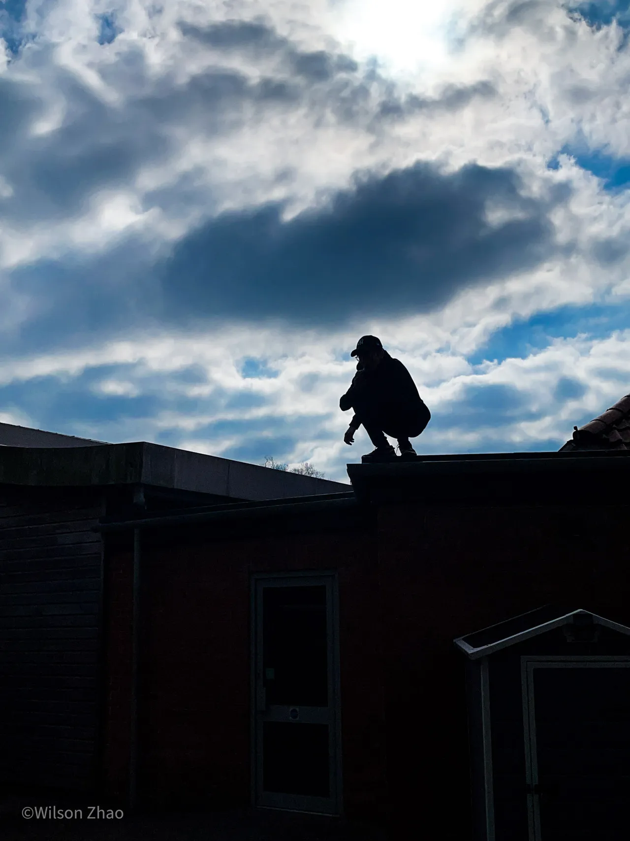 A figure crouches in silhouette on the edge of a rooftop against a bright, broken sky.