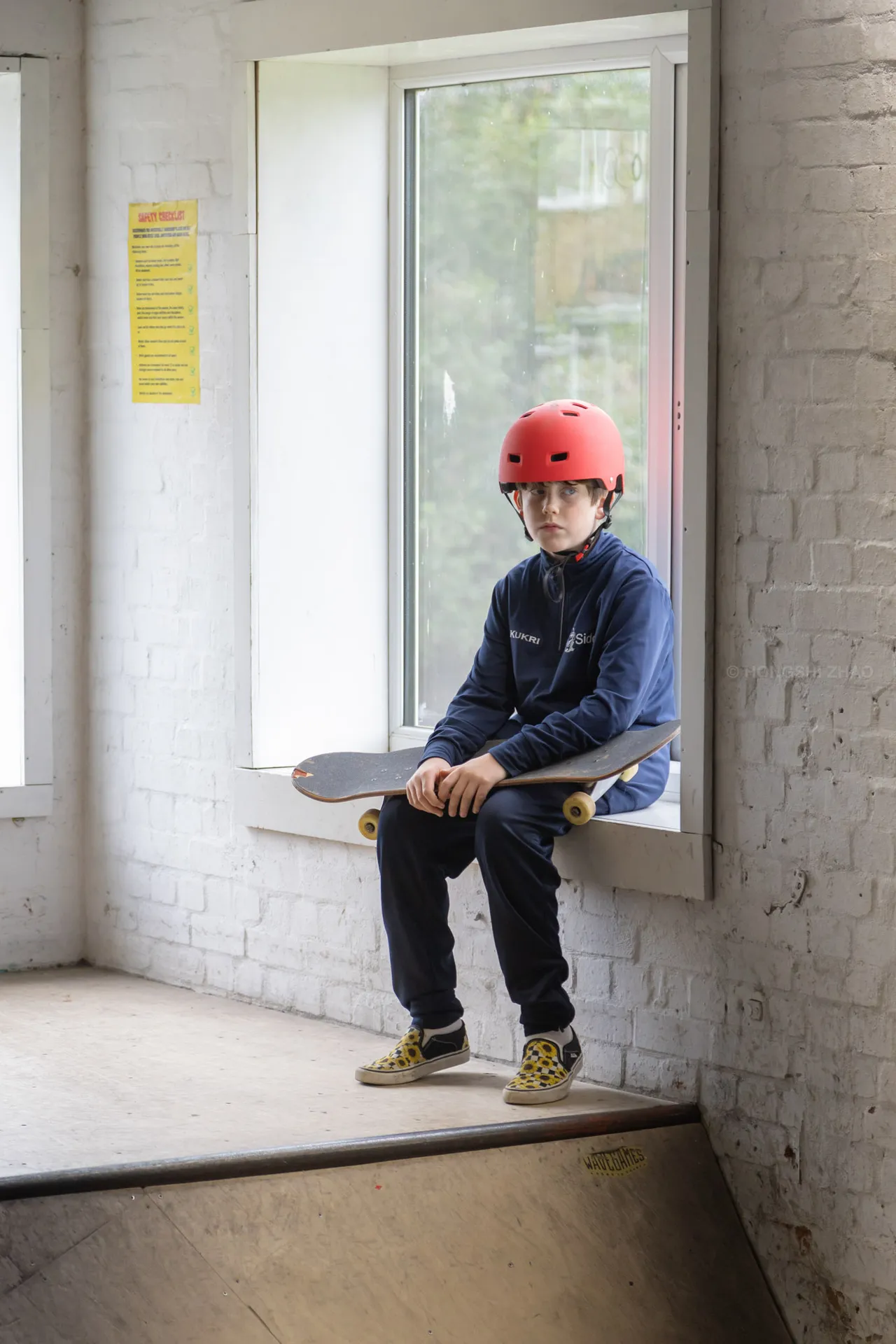 A boy in a red helmet sits on a whitewashed window ledge with a skateboard beside him.