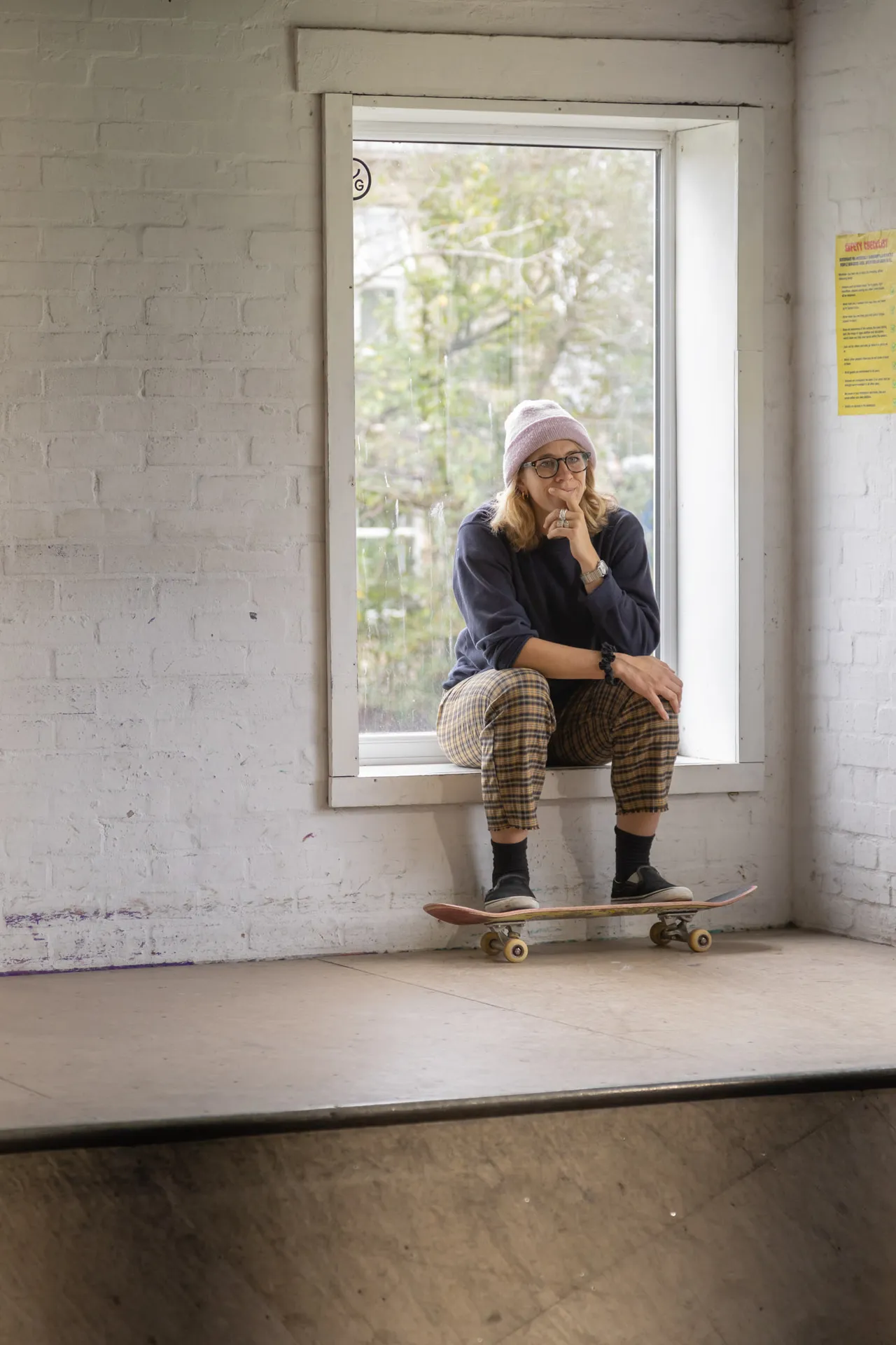 A young man in a beanie sits on a window ledge above a skate ramp, feet resting on his board.