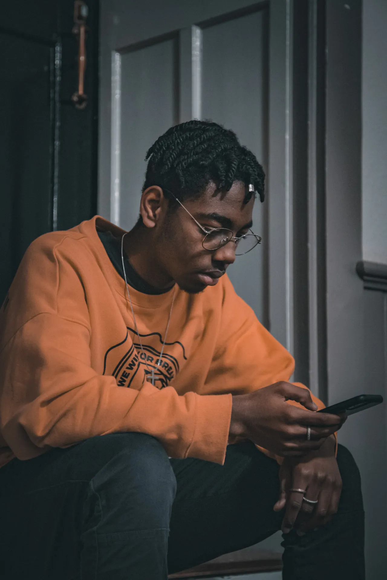 A young man in an orange sweatshirt and glasses sits in a dim doorway, looking down at his phone.