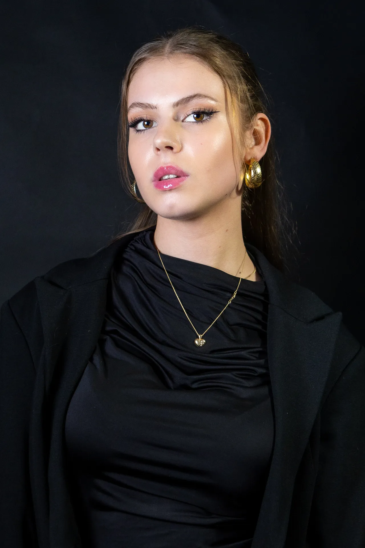 Studio portrait of a young woman dressed in black with gold hoop earrings, against a dark backdrop.