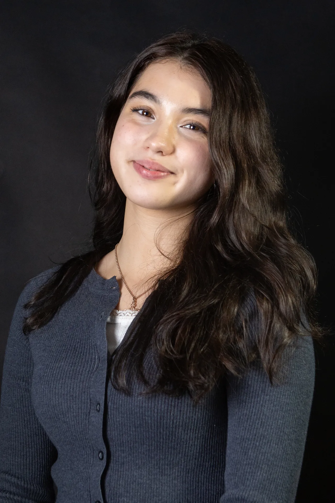 Studio portrait of a smiling young woman with long dark hair in a grey cardigan, against a dark backdrop.