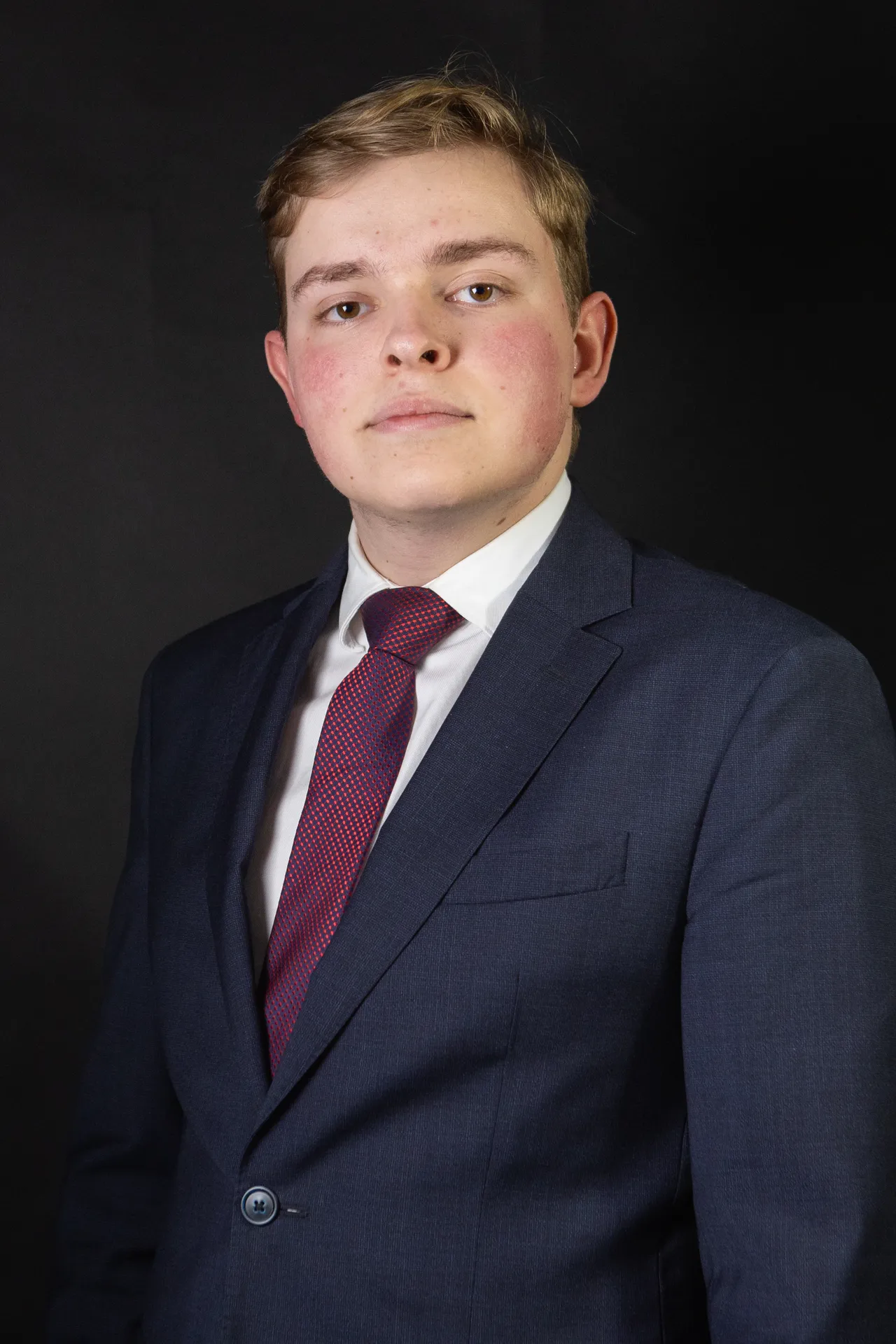 Studio portrait of a young man in a navy suit and burgundy tie, facing the camera against a dark backdrop.