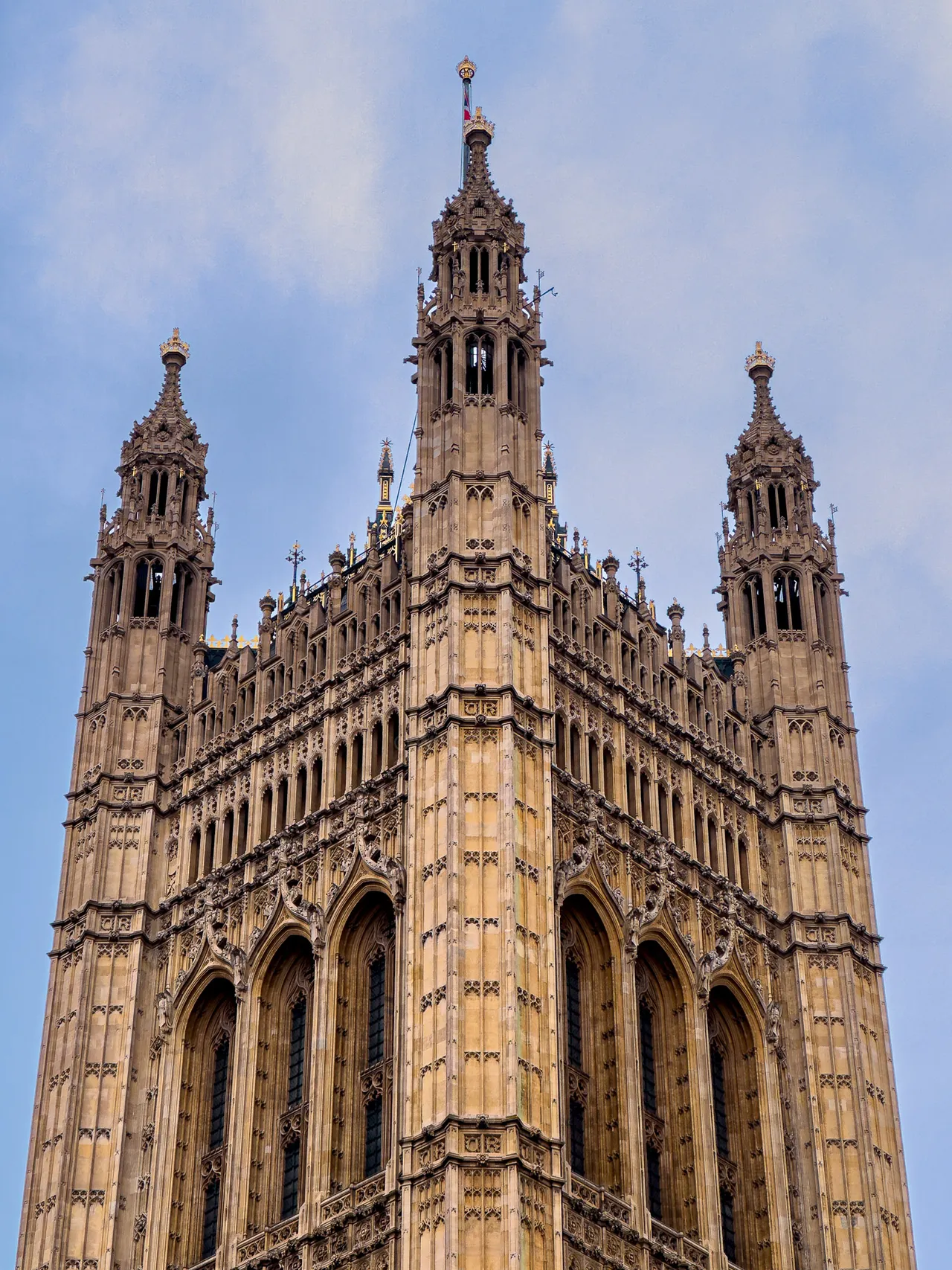 Looking up at an ornate Gothic stone tower against a pale blue sky.
