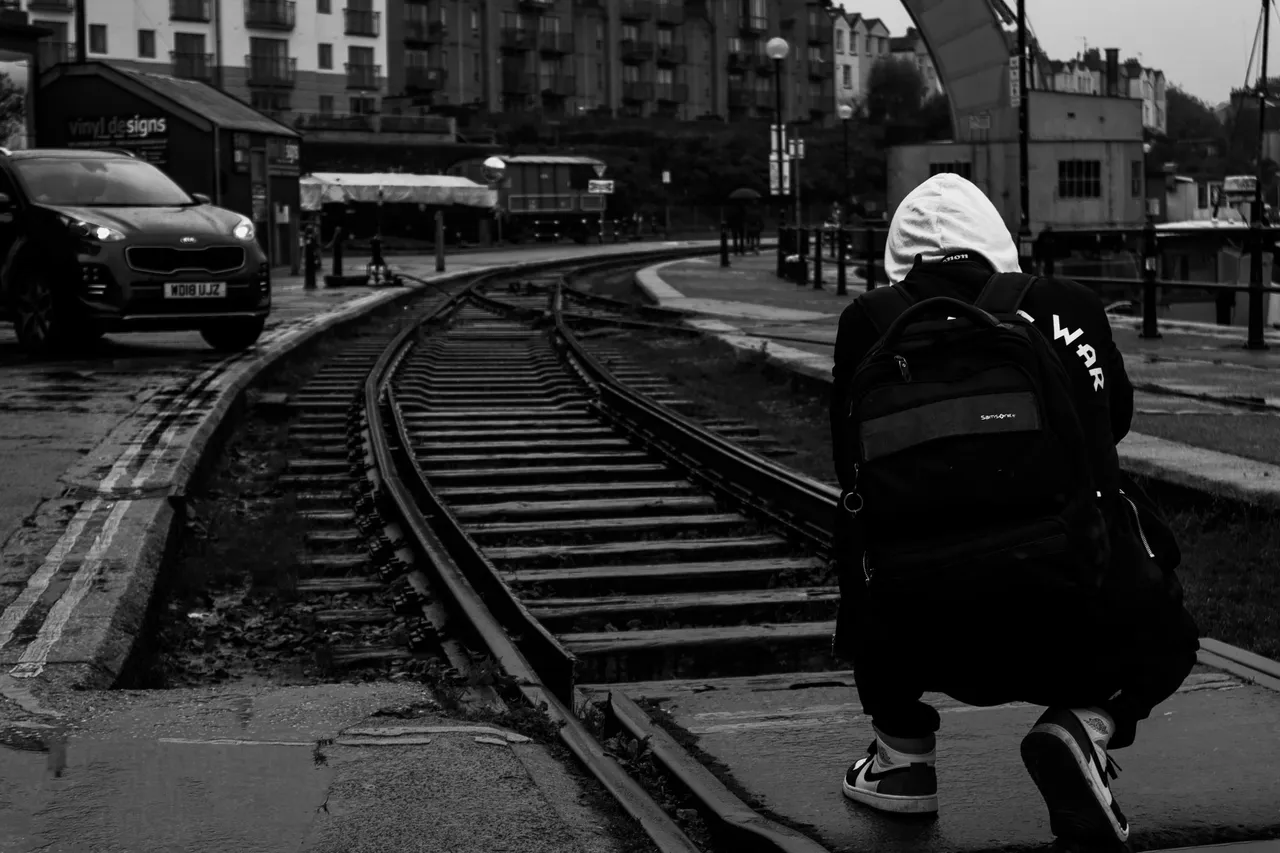 In black and white, a hooded figure with a backpack crouches beside old railway tracks set into a harbourside street.