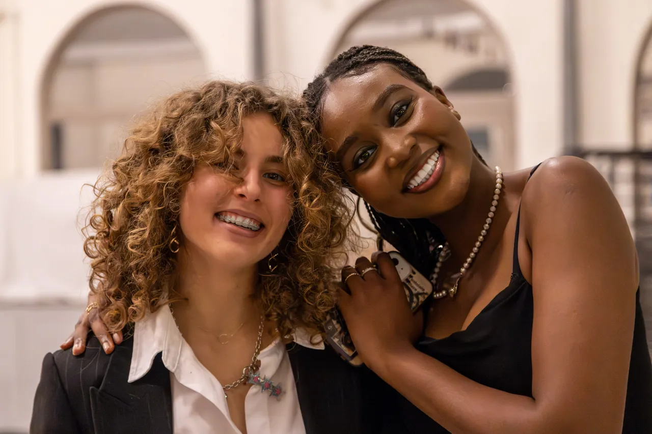 Two friends lean into each other and laugh — one with curly hair and a black blazer, the other in a black slip dress and pearls.