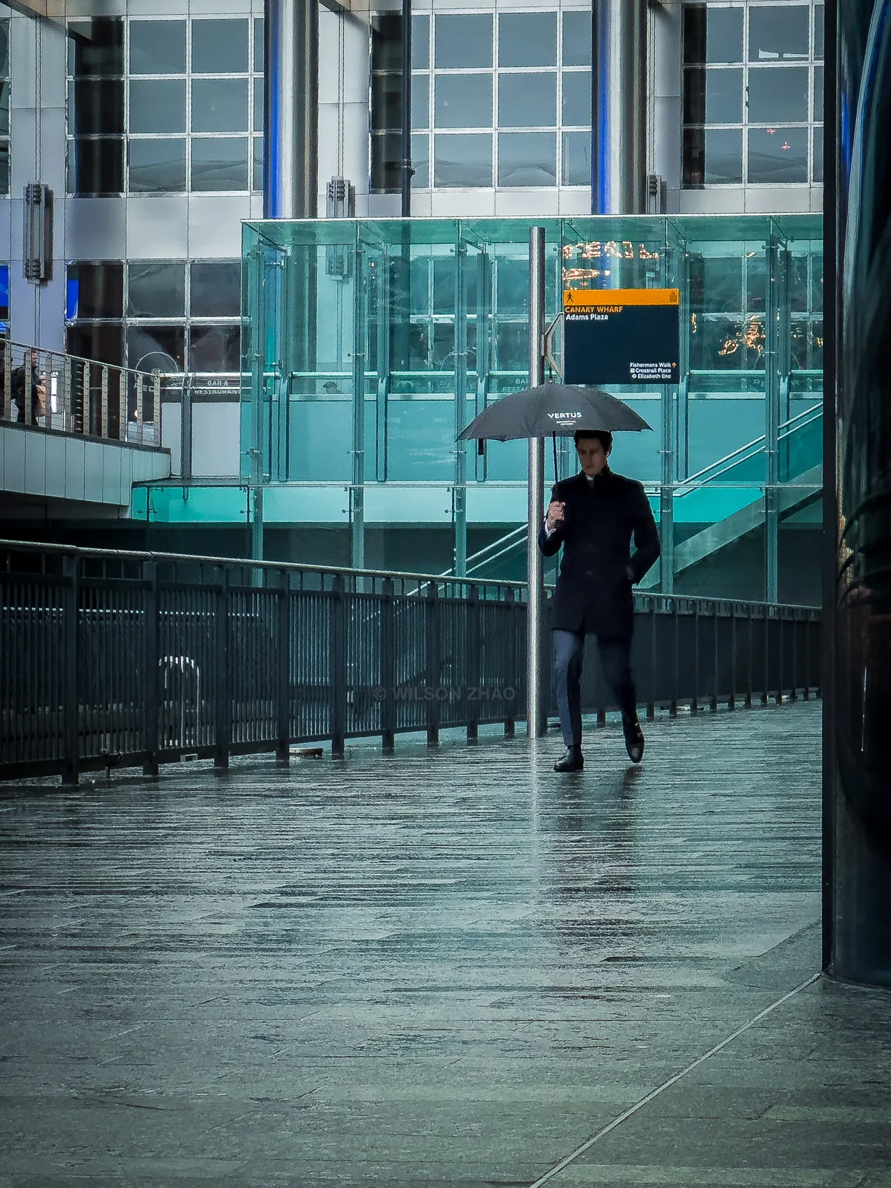 A man with an umbrella walks along a rain-slicked walkway below a glass building lit in teal.