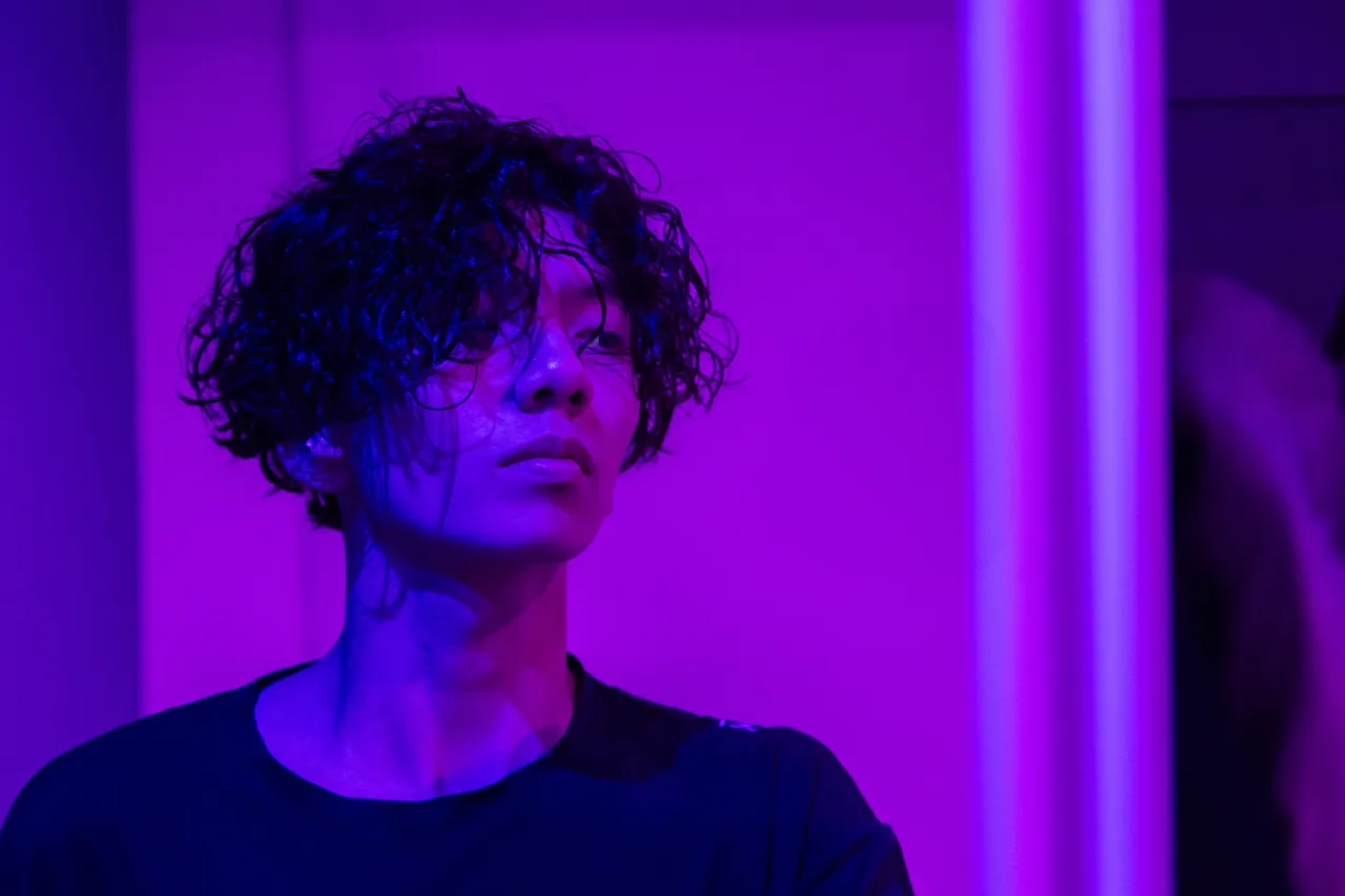A young man with curly hair lit in saturated violet light, looking up and away from the camera.