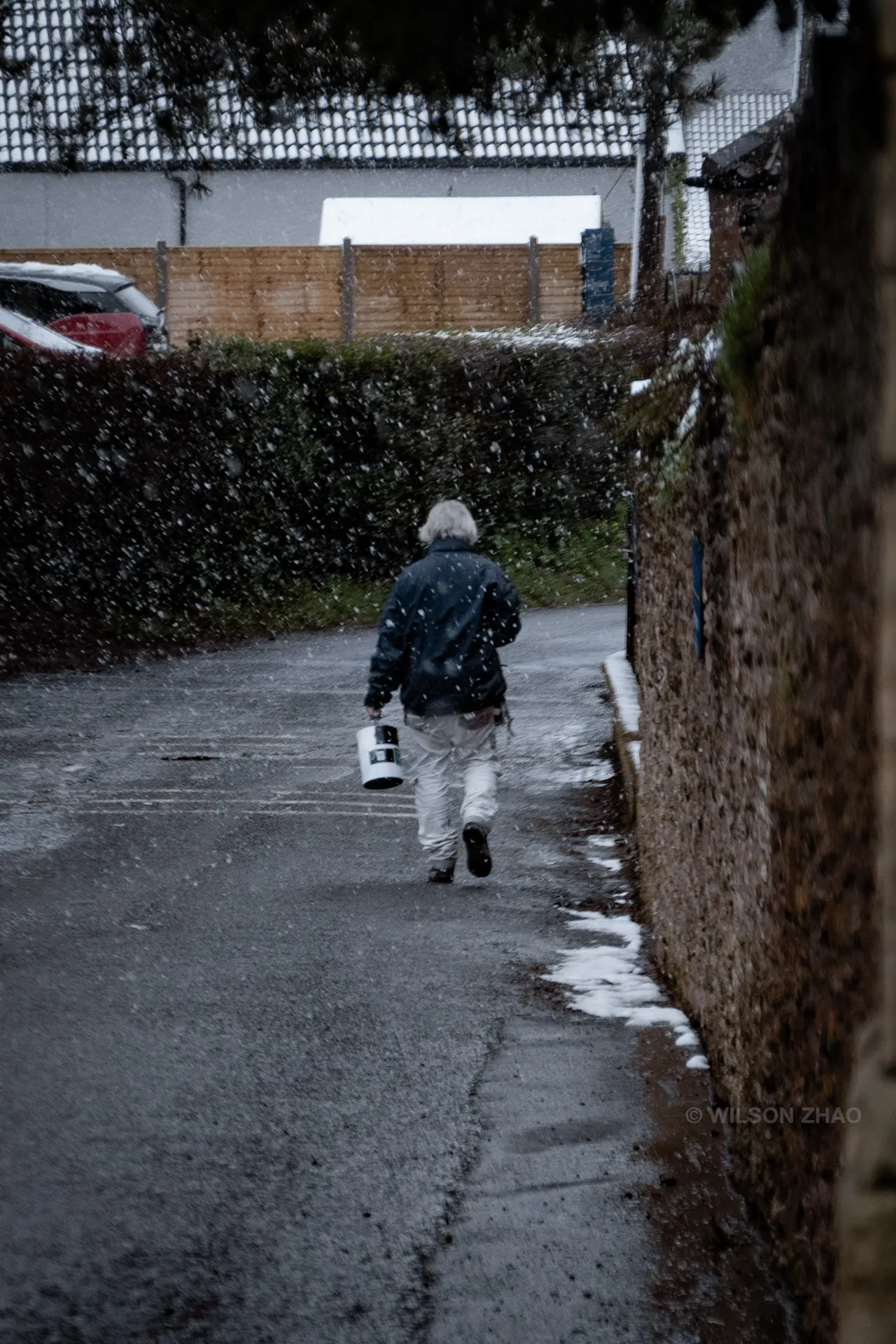 An elderly man walks away down a narrow lane between a hedge and a stone wall, patches of snow at the kerb.