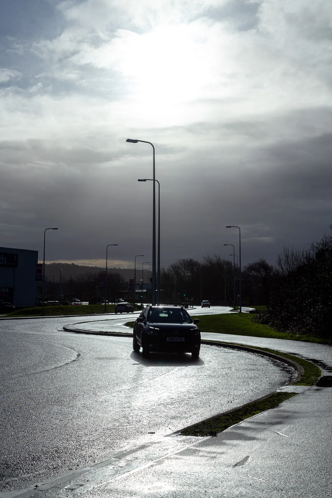 A single car rounds a curve on a wet road, backlit by low sun breaking through heavy cloud.
