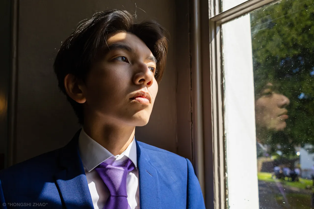 A young man in a blue suit and lilac tie stands by a window, side-lit by low sun, his reflection faint in the glass.